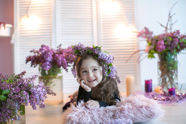 Girl child with a wreath of lilac on his head sits on the floor surrounded by a vase of flowers - Valokuva, kuva