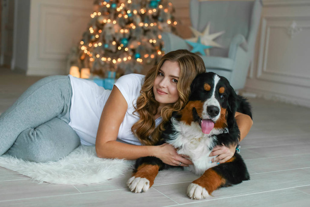 A beautiful woman with long hair lies on a rug against the background of a Christmas tree and next to her lies her big dog - Fotoğraf, Görsel