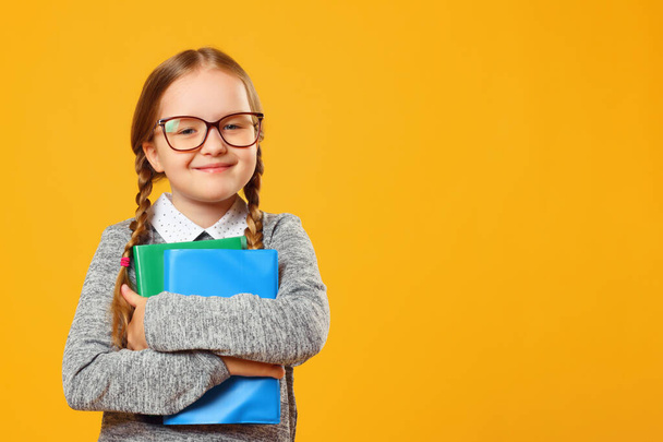 Cheerful little girl schoolgirl in glasses holding books on a yellow background wall. The concept of education and school. Copy space - Photo, Image