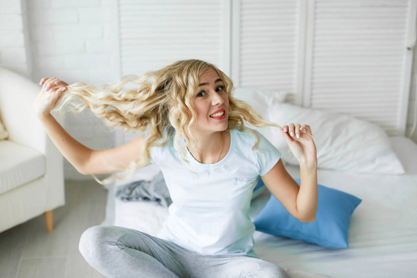 The girl sits on the bed and touches her curly blonde long hair - Photo, image