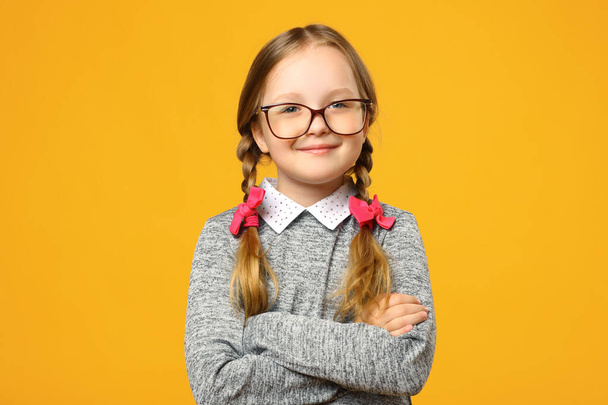 Closeup portrait of a funny little girl in glasses on a yellow background. Child schoolgirl folded her arms and looks into the camera - 写真・画像