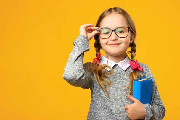 Portrait of a cute little kid girl on a yellow background. Child schoolgirl looking at the camera, holding a book and straightens glasses. The concept of education. Copy space - Photo, image