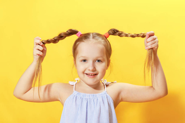 Closeup portrait of a cheerful little girl on a yellow background. The child looks into the camera and holds his hands pigtails out of hair - Fotografie, Obrázek
