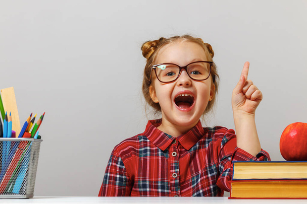 Funny little preschool girl in glasses sits at the table with books and points finger up. The concept of education. Gray background - Φωτογραφία, εικόνα