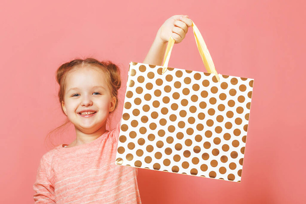 Closeup portrait of a cute little girl with hair buns over pink background. The child lifted the paper bag high, rejoicing at the shopping or gift - Zdjęcie, obraz