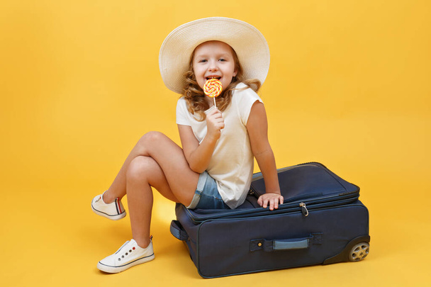 Malá blonďatá holčička sní o cestování a sedí na kufříku s bonbóny v ruce. Žluté pozadí - Fotografie, Obrázek