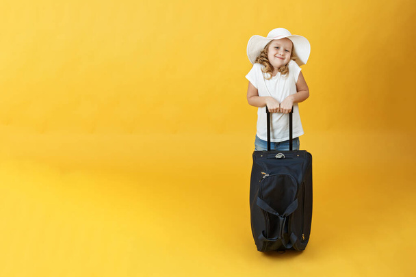 Mała dziewczynka blondynka jest gotowa do podróży i stoi z walizką. Żółte tło - Zdjęcie, obraz