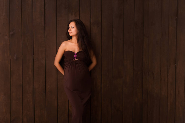 Mujer embarazada en vestido rojo toma las manos en el vientre sobre un fondo marrón oscuro. Embarazo, maternidad, concepto de expectativa. Hermosa foto del estado de ánimo tierno del embarazo
 - Foto, imagen