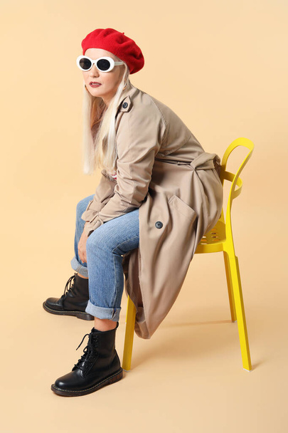 Fashionable mature woman sitting on chair against color background - Photo, Image