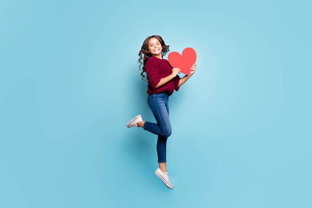 Full length body size side profile photo of schoolgril jumping up holding big red heart smiling toothily in burgundy sweater receiving present gift isolated blue vivid color background - Фото, изображение