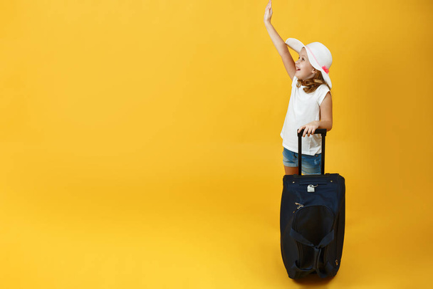 Little child girl blonde ready for the journey, standing with a suitcase and hand outstretched up. Yellow background - Foto, imagen