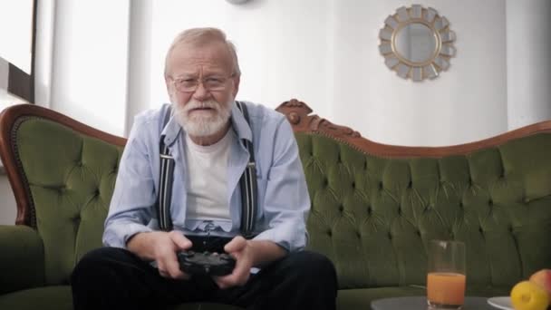 modern grandfather gamer with beard and glasses for vision plays video games sitting on couch and holding joystick in hands - Footage, Video