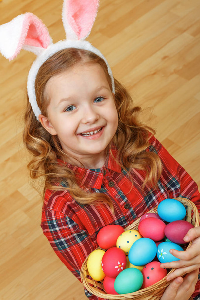A cute little child girl with bunny ears sits on the floor with a basket of Easter eggs - Fotoğraf, Görsel