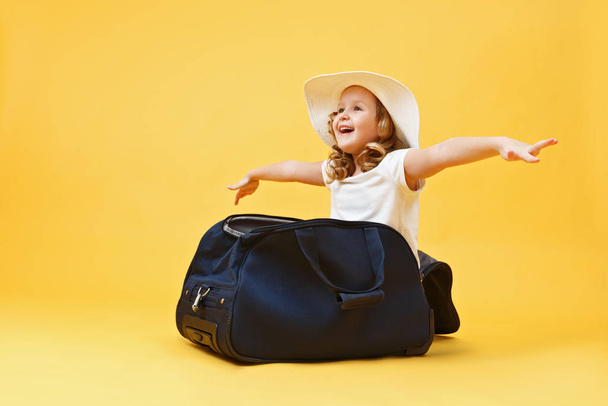 Όνειρα ταξιδιού. Ένα μικρό κοριτσάκι κάθεται σε μια βαλίτσα και παίζει τον πιλότο του αεροπλάνου και τον ταξιδιώτη. Κίτρινο φόντο - Φωτογραφία, εικόνα