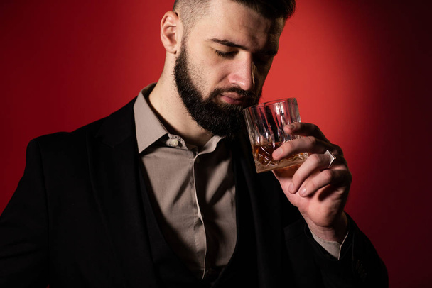 Schöner bärtiger Mann im Anzug mit stylischem Bart, der ein Glas Whisky in der Hand hält und es auf rotem Grund trinken wird. Nahaufnahme - Foto, Bild