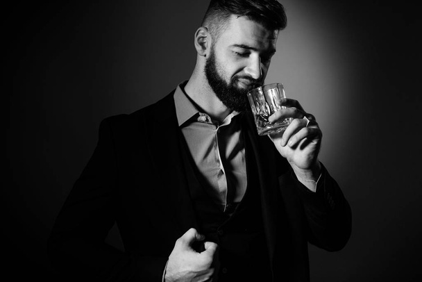 Красивый бородатый мужчина в костюме со стильной бородой, держащий в руках стакан виски и собирающийся его выпить, на красном фоне. Черное и белое - Фото, изображение