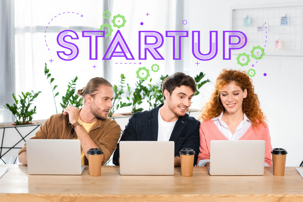 tre amici sorridenti seduti a tavola e utilizzando computer portatili in ufficio con illustrazione startup
 - Foto, immagini
