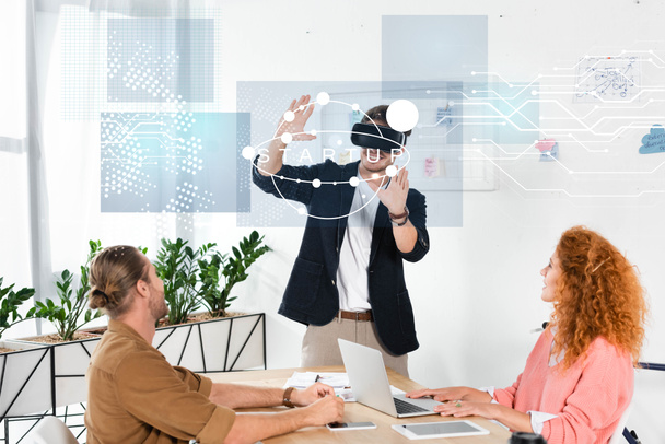 smiling businessman with vr headset gesturing in office near colleagues and startup illustration - Photo, Image