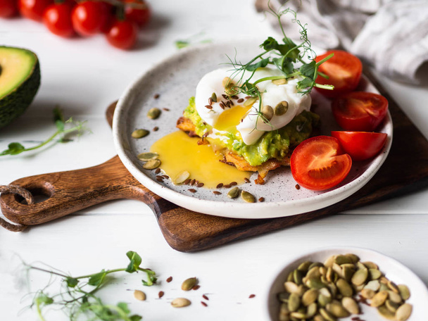 gesundes, nahrhaftes Frühstück mit Kartoffelpuffer, pürierter Avocado, pochiertem Ei, Erbsensprossen und verschiedenen Samen auf einem Keramikteller - Foto, Bild
