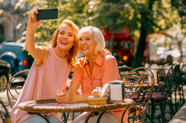 Cheerful aged woman sitting at cafe terrace with daughter and posing for selfie - Foto, Imagen