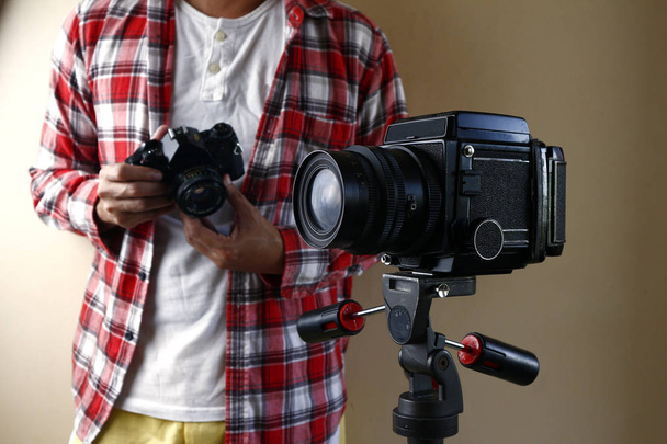 Adam 35 mm 'lik bir kamera tutuyor ve tripodun üzerinde orta formatlı bir film kamerasının arkasında duruyor. - Fotoğraf, Görsel