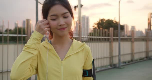 Beautiful young Asia athlete lady exercises using smartphone for listen to music while running in urban environment. Korean teen girl wearing sports clothes on walkway bridge in early morning. - Footage, Video