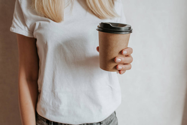 Woman holding in hand paper cup with coffee or tea. Drink coffee time. Disposable paper cup. Beautiful blond hair young girl. Casual style, white shirt and jeans. Take away. Hot beverage. Blank space - Photo, image