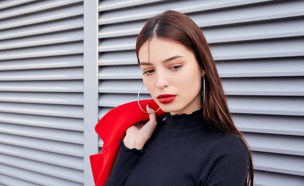 Calm pretty girl with puffy red lips standing on gray striped wallback, throwing red overcoat over her shoulder - Fotografie, Obrázek