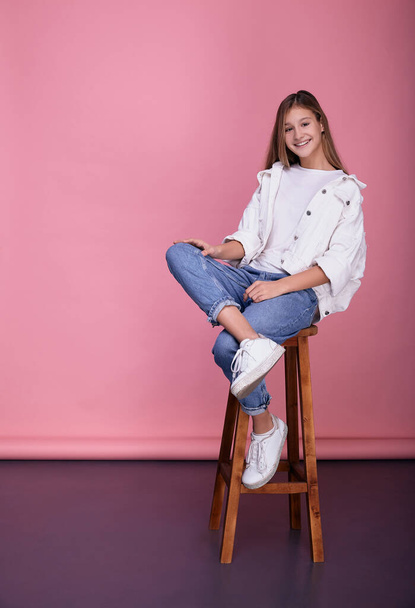 Fashionably dressed young girl sits on a chair on a pink background. - Foto, Bild