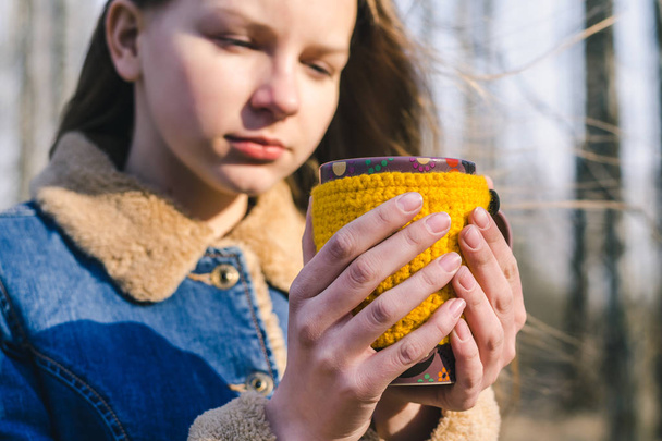 junges schönes Mädchen mit langen Haaren hält im Park an einem klaren, sonnigen, kalten Tag einen Becher in einer gelben, selbstgestrickten Hülle mit einem heißen Getränk in den gefrorenen Händen - Foto, Bild