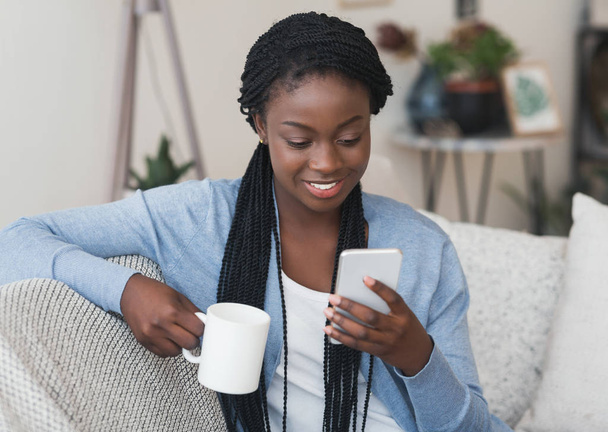 Nuori afrikkalainen amerikkalainen tyttö lukee viestiä älypuhelimella ja juo kahvia
 - Valokuva, kuva