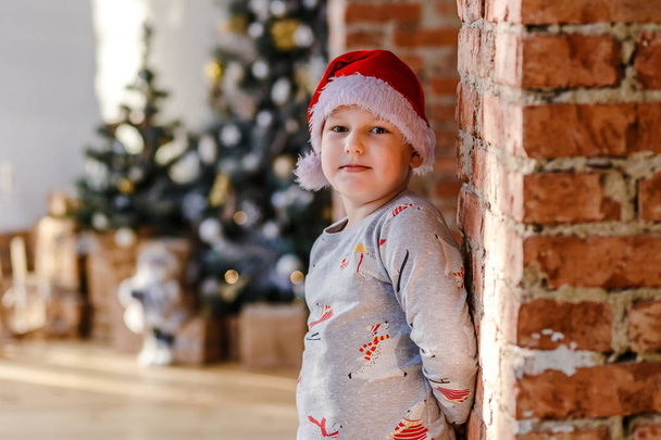 an eight year old boy in pajamas and a Santa hat stands near a brick wall against the background of a Christmas tree - Foto, Imagen