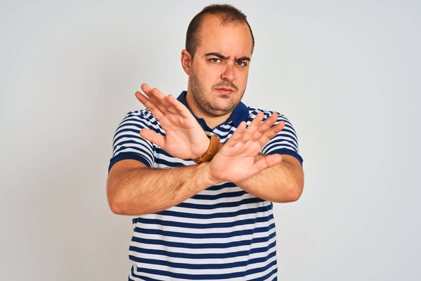 Молодий чоловік у повсякденному смугастому поло, що стоїть над ізольованим білим тлом Вираз відторгнення, що перетинає руки та долоні, роблять негативний знак, сердите обличчя
 - Фото, зображення