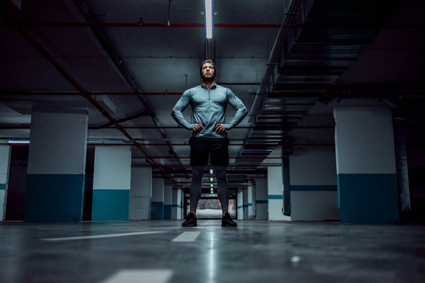 Низкий угол обзора привлекательного кавказского спортсмена в активной одежде, стоящего в подземном гараже с руками на бедрах. Концепция городской жизни
. - Фото, изображение