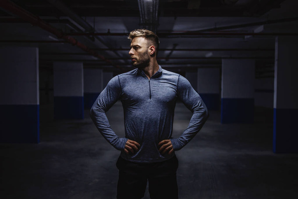 Powerful strong caucasian man in sportswear standing in garage with hands on hips and looking away. Nighttime. Urban life concept. - Photo, Image