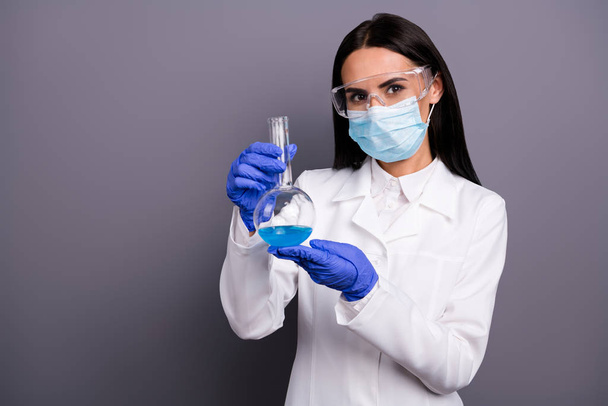 Portrait of successful scientist woman present her new medication against cancer she hold in her flask blue liquid wear safety gloves goggles white coat mask isolated grey color background - Foto, Imagem