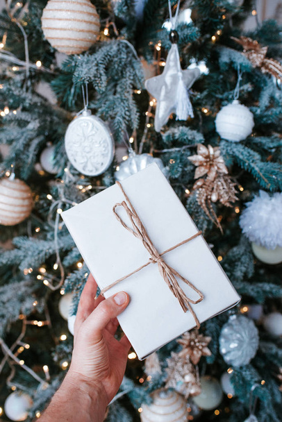 Man holds a Christmas present in hands. Happy holiday, gift box. Christmas tree with garlands in home, beautiful lights. Surprise for Boxing day, New Year and Christmas. Kraft wrapping paper and twine - Φωτογραφία, εικόνα