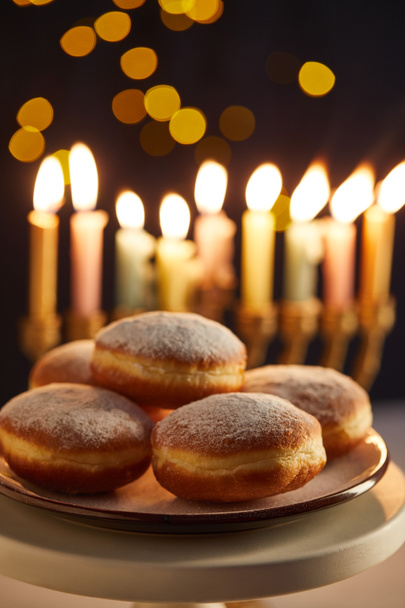 selective focus of delicious doughnuts on stand near glowing candles on black background with bokeh lights on Hanukkah - Fotó, kép