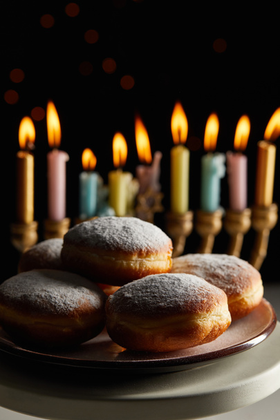 selective focus of delicious doughnuts on stand near glowing candles in menorah on black background with bokeh lights on Hanukkah - Photo, Image