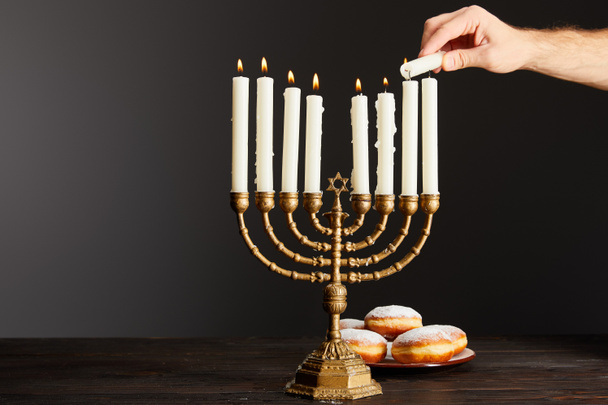 cropped view of man lighting up candles in menorah near doughnuts on black background on Hanukkah - Fotó, kép