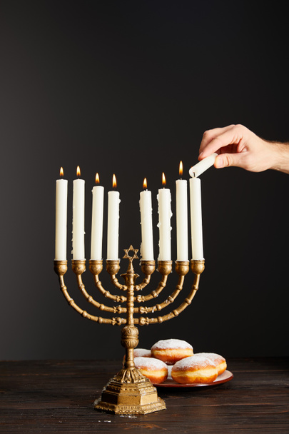 cropped view of man lighting up candles in menorah near doughnuts on black background on Hanukkah - Photo, Image