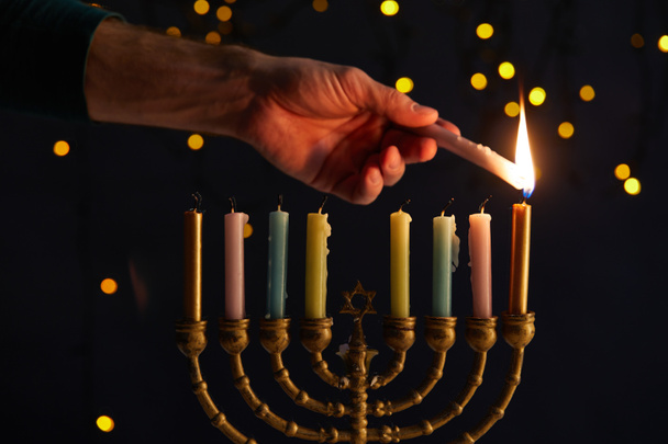 gedeeltelijke weergave van de mens het aansteken van kaarsen in menorah op zwarte achtergrond met bokeh lichten op Chanoeka - Foto, afbeelding