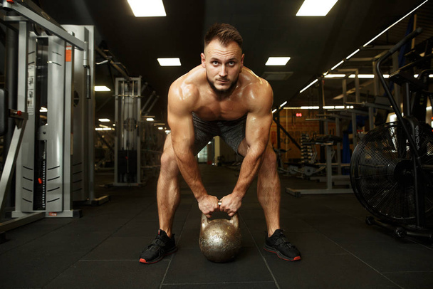 Strong handsome bearded man holding kettlebell and doing dumbbell front swings in the popular dark gym. SPORT IS LOVE. - Photo, Image