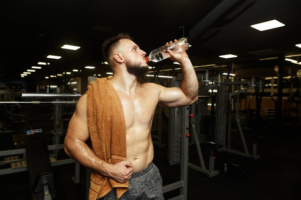 Αθλητικός όμορφος άντρας πίνει νερό μετά τη βροχή στο άδειο γυμναστήριο. - Φωτογραφία, εικόνα