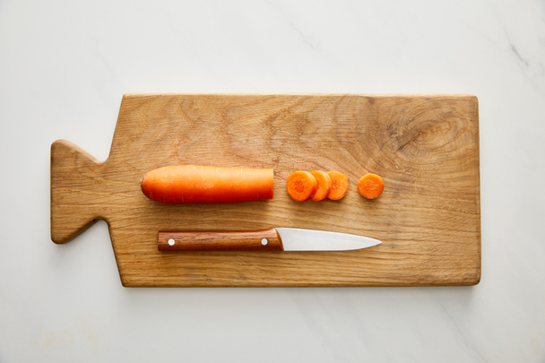 Top view of carrot with slices and knife on cutting board on marble background - Photo, image