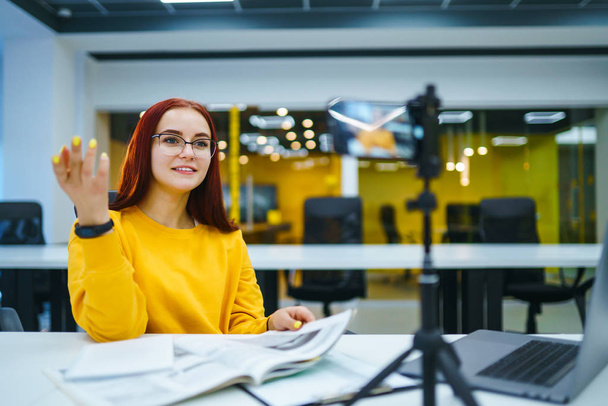 Female blogger with laptop recording video while sitting at office and talking  about startup small business. Teenager student in a yellow sweater having fun vlogging live feeds on social media. - Photo, Image