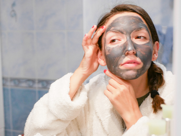 Το κορίτσι φροντίζει το πρόσωπο. Καλλυντική μαύρη μάσκα. Μια γυναίκα φέρνει ομορφιά στο πρόσωπό της, ξεφορτώνεται τις μαύρες κουκίδες με μια μαύρη μάσκα. Η γυναίκα στο μπάνιο κοιτά στον καθρέφτη. - Φωτογραφία, εικόνα