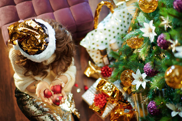 Upper view of stylish middle age housewife with long brunette hair in gold sequin skirt and white sweater under decorated Christmas tree near present boxes holding broken Christmas ball. - Foto, Imagen