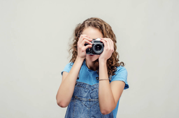 Curly brunette girl in denim overalls is holding a vintage film camera in her hands. Takes a picture - Foto, imagen