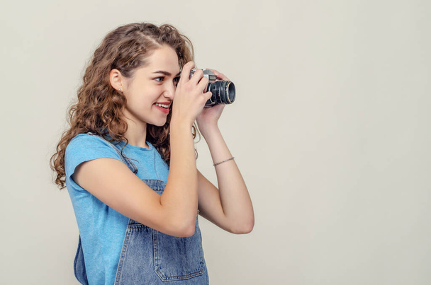 Fille brune bouclée en salopette de denim tient une caméra de film vintage dans ses mains. Il prend une photo. Vue latérale
. - Photo, image